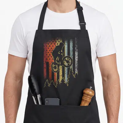 Mountain biking bike apron - Distressed American flag with mountain biker silhouette and tree line in beige red blue — shown on a black cotton canvas apron against a white background