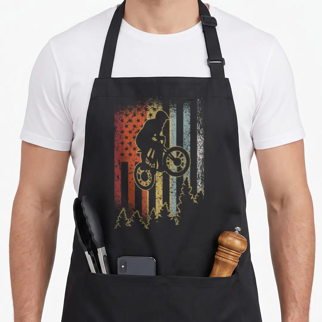 Mountain biking bike apron - Distressed American flag with mountain biker silhouette and tree line in beige red blue — shown on a black cotton canvas apron against a white background