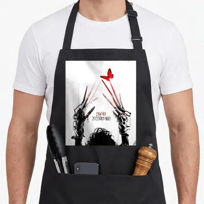 Tim butterfly apron - Edward Scissorhands text, two raised scissor hands, red butterfly, top of head hair — shown on a black cotton canvas apron against a white background
