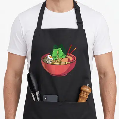 Eating frog aprons - Green cartoon frog sitting in red ramen bowl with seaweed and chopsticks — shown on a black cotton canvas apron against a white background