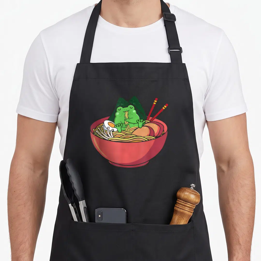 Eating frog aprons - Green cartoon frog sitting in red ramen bowl with seaweed and chopsticks — shown on a black cotton canvas apron against a white background