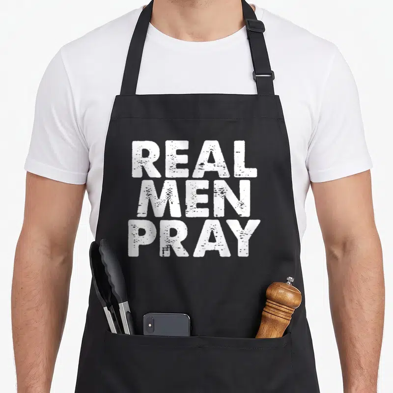 Pray catholic apron - White distressed text reading Real Men Pray stacked in three lines — shown on a black cotton canvas apron against a white background
