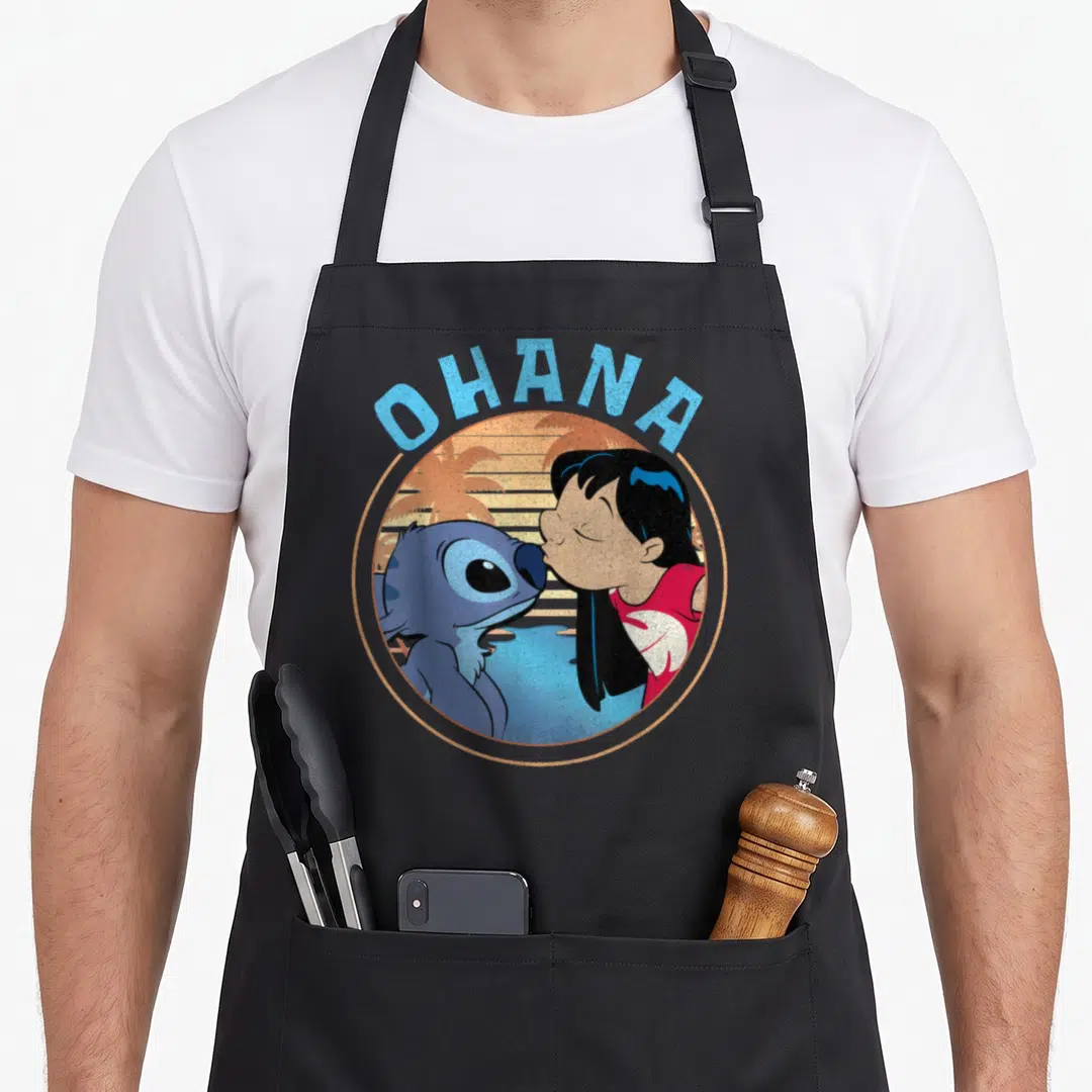 Ohana novelty apron - Lilo and Stitch kissing in circle with OHANA text and palm trees — shown on a black cotton canvas apron against a white background
