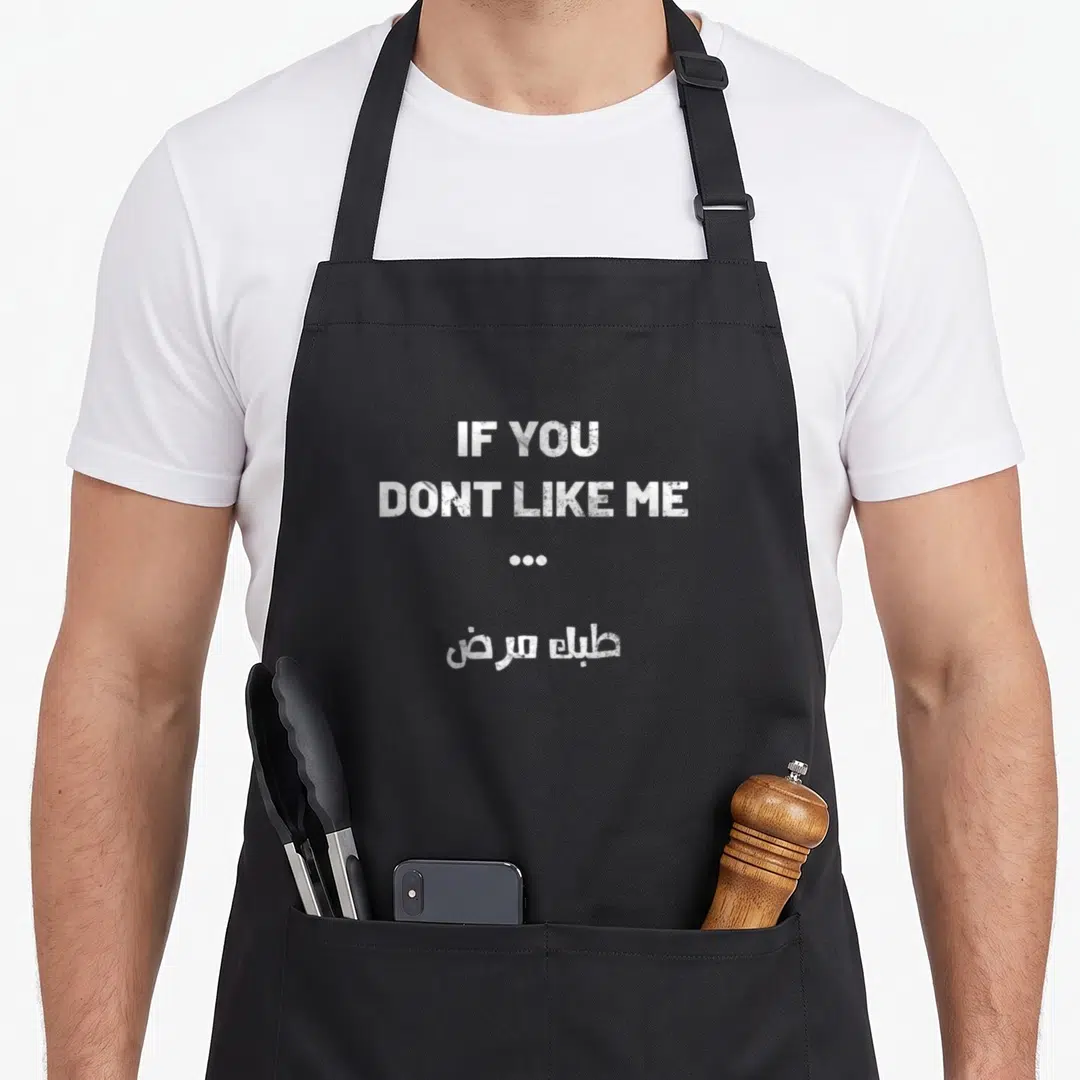 Iraqi joke apron - White stacked text reading IF YOU DONT LIKE ME followed by Arabic script meaning it is your disease — shown on a black cotton canvas apron against a white background