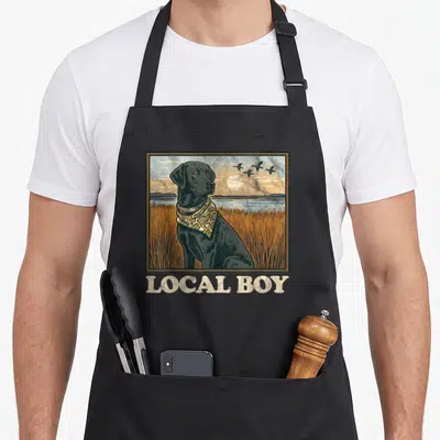 Local dog apron - Framed black lab in orange marsh with birds and LOCAL BOY text below — shown on a black cotton canvas apron against a white background