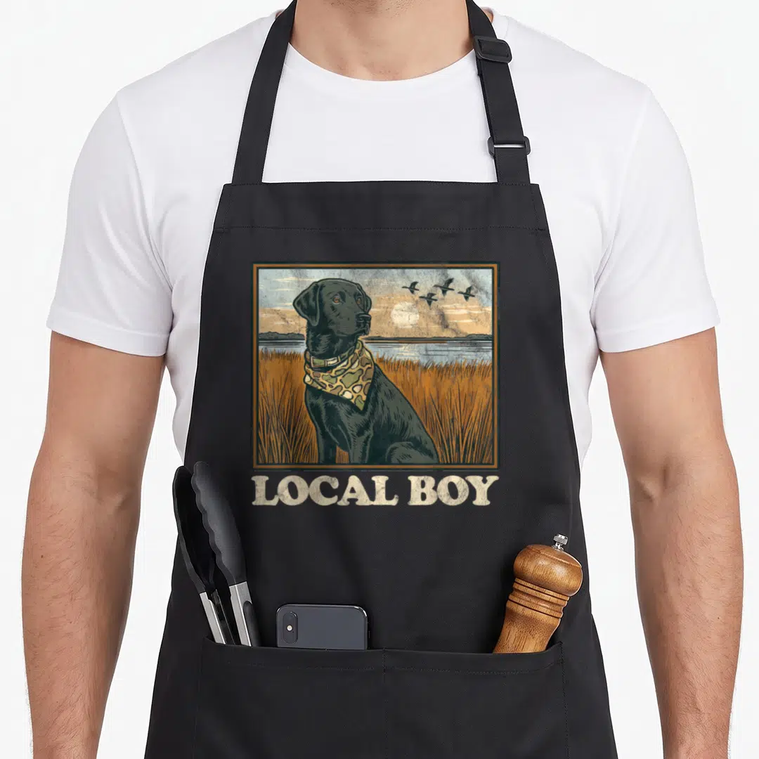 Local dog apron - Framed black lab in orange marsh with birds and LOCAL BOY text below — shown on a black cotton canvas apron against a white background
