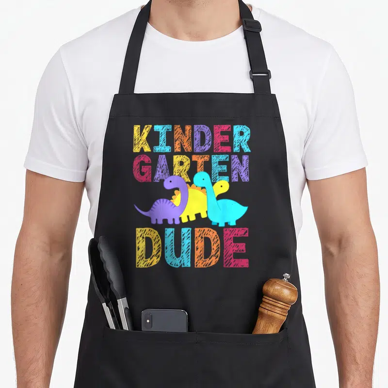 Kindergarten school dude apron - Stacked text KINDER GARTEN DUDE surrounding three cartoon dinosaurs in yellow, blue — shown on a black cotton canvas apron against a white background