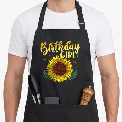 Birthday sunflower apron - Stacked Birthday GIRL text above a large yellow sunflower with green leaves and sparkles — shown on a black cotton canvas apron against a white background