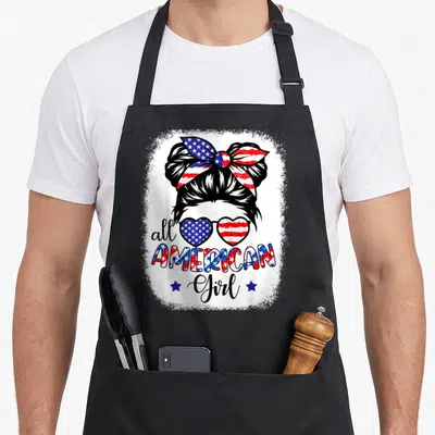 Patriot 4th of july apron - All American Girl text with flag-patterned sunglasses and bun silhouette flanked by two — shown on a black cotton canvas apron against a white background