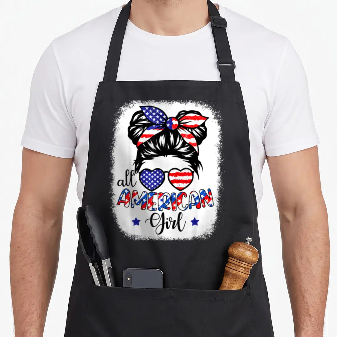 Patriot 4th of july apron - All American Girl text with flag-patterned sunglasses and bun silhouette flanked by two — shown on a black cotton canvas apron against a white background