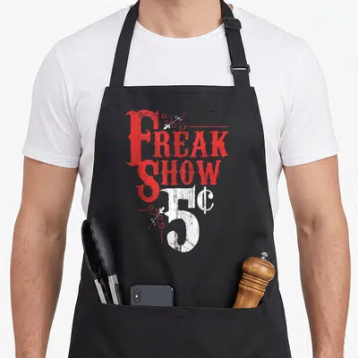 Freak weird aprons - Vertical red and white text reading FREAK SHOW 5¢ with distressed swirls — shown on a black cotton canvas apron against a white background