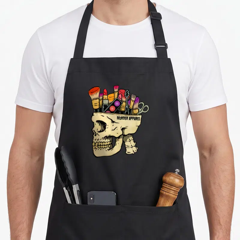 Addict gothic apron - Cartoon skull filled with makeup brushes and palettes reading MURDER APPAREL — shown on a black cotton canvas apron against a white background