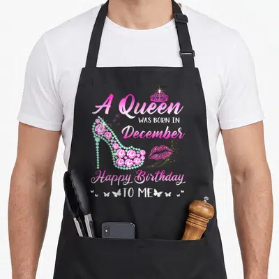 Vintage queen december birthday apron - Pink script text with jeweled high heel, glittery lips and white butterflies — shown on a black cotton canvas apron against a white background