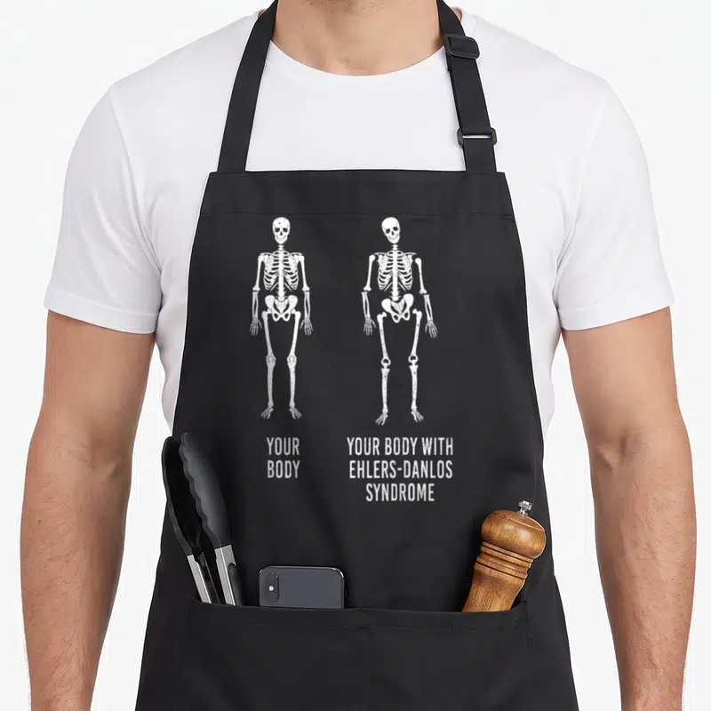 Ehler body apron - Two white skeletons comparing normal anatomy to Ehlers-Danlos Syndrome with stacked distressed text — shown on a black cotton canvas apron against a white background