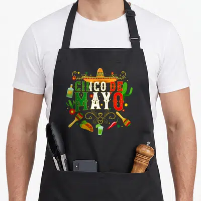 Fiesta taco apron - Cinco De Mayo text with sombrero, maracas, taco, chili, and cactus in green white red yellow — shown on a black cotton canvas apron against a white background