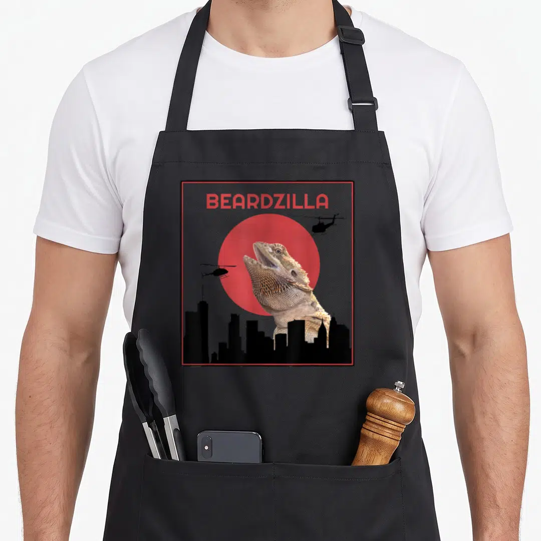Beardzilla dragon apron - Bearded dragon photo with BEARDZILLA text, red circle, helicopters, and city skyline — shown on a black cotton canvas apron against a white background