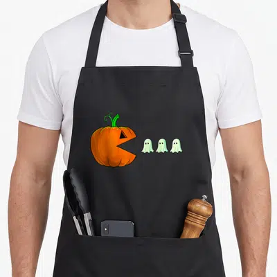 Pumpkin ghost halloween apron - Orange cartoon pumpkin with open mouth facing three small white ghosts in a row — shown on a black cotton canvas apron against a white background