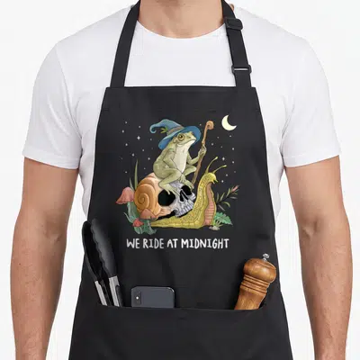 Witch cottagecore apron - Frog in blue hat on skull snail with staff, WE RIDE AT MIDNIGHT text, pink mushrooms, stars — shown on a black cotton canvas apron against a white background