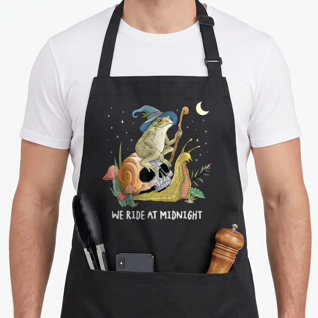 Witch cottagecore apron - Frog in blue hat on skull snail with staff, WE RIDE AT MIDNIGHT text, pink mushrooms, stars — shown on a black cotton canvas apron against a white background