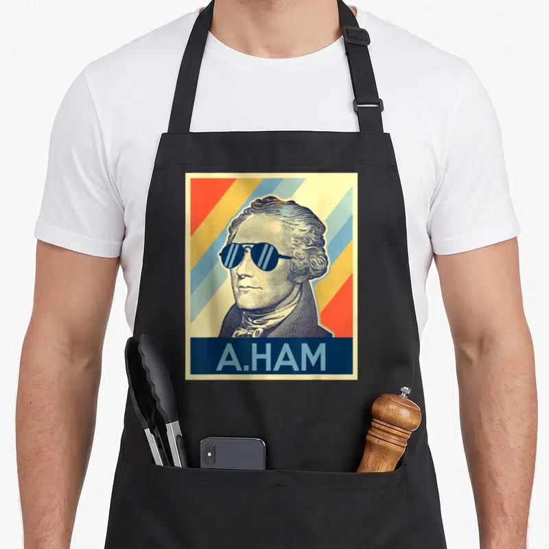 Hamilton musicals apron - Alexander Hamilton portrait with sunglasses and A.HAM text on red yellow blue cream stripes — shown on a black cotton canvas apron against a white background