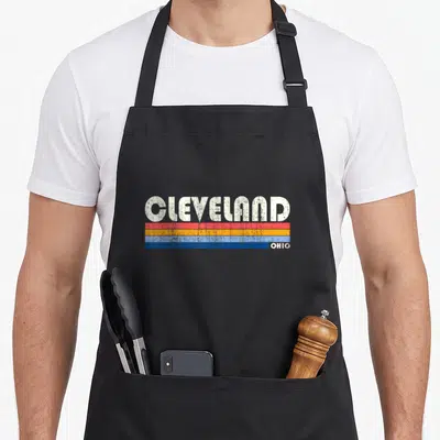 Cleveland 70s apron - CLEVELAND text over red orange blue stripes with OHIO label on right — shown on a black cotton canvas apron against a white background