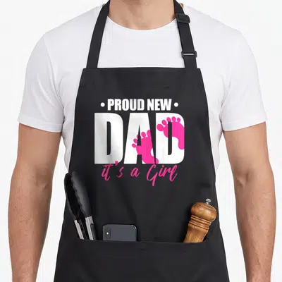 Parent crew apron - White PROUD NEW DAD text with two pink baby footprints and pink script reading it's a Girl — shown on a black cotton canvas apron against a white background