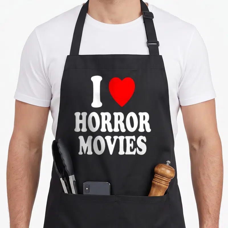 Scary horror apron - White I and red heart above stacked Horror Movies text — shown on a black cotton canvas apron against a white background