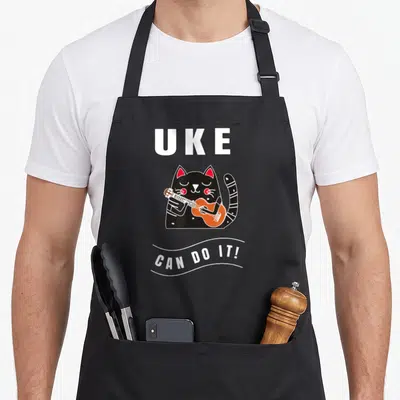 Ukulele cat apron - Cartoon black cat playing orange uke with text UKE CAN DO IT! — shown on a black cotton canvas apron against a white background