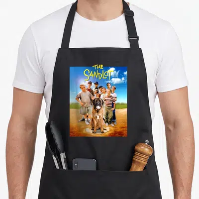 Sandlot 90s apron - Stacked yellow title above photo of boys and dog on baseball diamond with blue sky — shown on a black cotton canvas apron against a white background