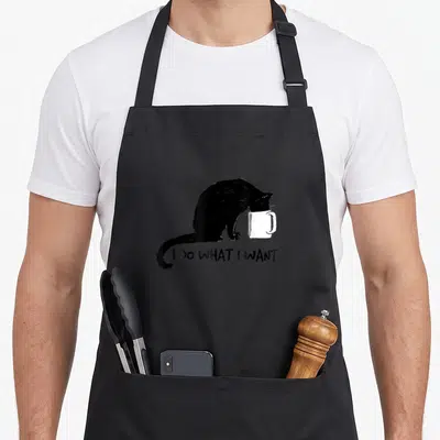 Cup cat apron - Black cat dipping head into white mug with text I DO WHAT I WANT below — shown on a black cotton canvas apron against a white background