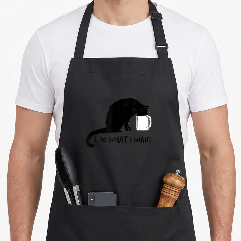 Cup cat apron - Black cat dipping head into white mug with text I DO WHAT I WANT below — shown on a black cotton canvas apron against a white background