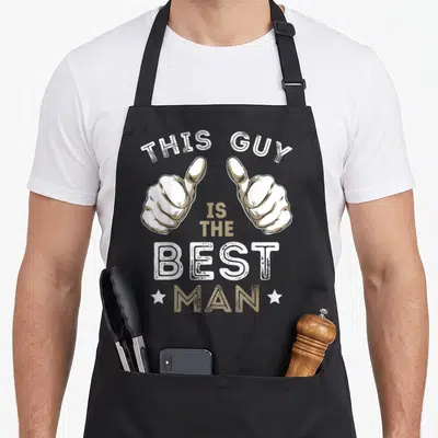 Funny bachelor funny wedding aprons - Two pointing hands in white and tan with distressed text reading This Guy Is — shown on a black cotton canvas apron against a white background