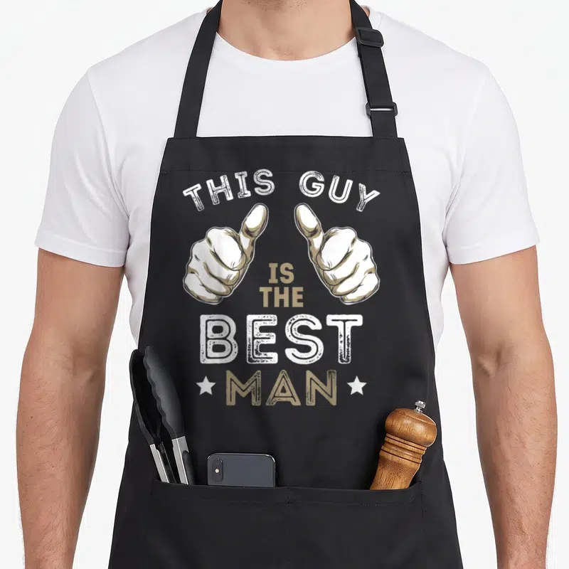 Funny bachelor funny wedding aprons - Two pointing hands in white and tan with distressed text reading This Guy Is — shown on a black cotton canvas apron against a white background