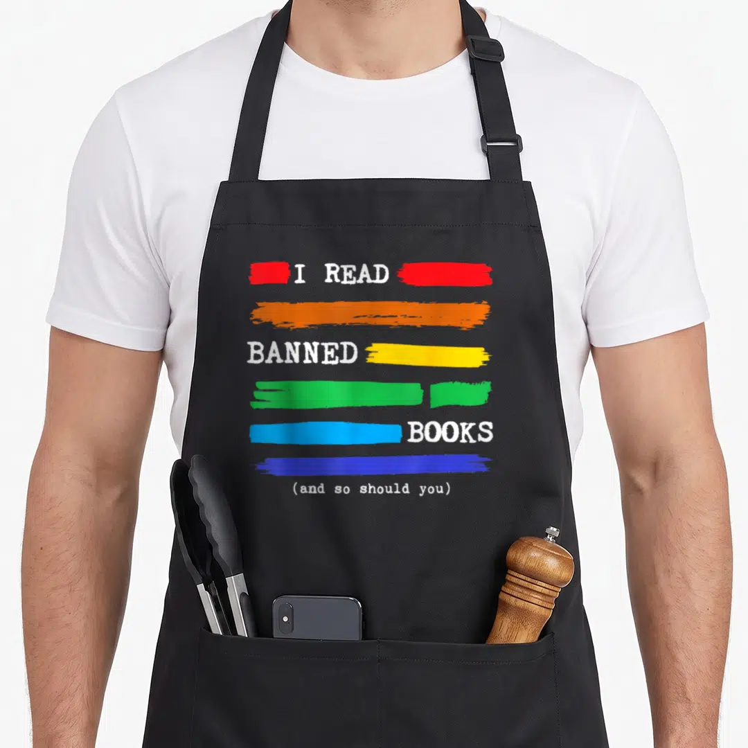 Read colorful aprons - White text I Read Banned Books with red orange yellow green blue horizontal brush strokes — shown on a black cotton canvas apron against a white background