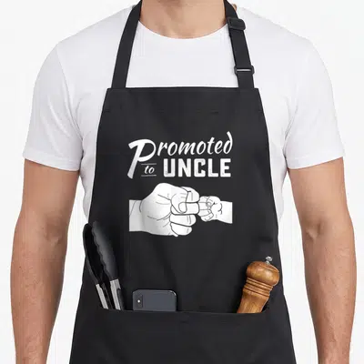Promoted uncle apron - White Promoted to Uncle text above adult and baby fistbump line art — shown on a black cotton canvas apron against a white background