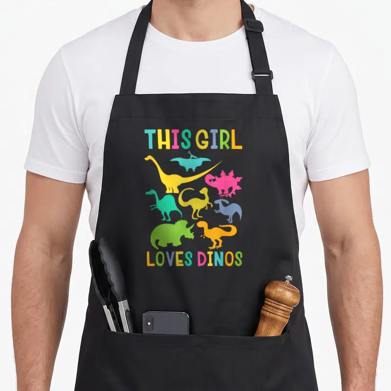 Cute dino dinosaur apron - Colorful dinosaur silhouettes surround THIS GIRL LOVES DINOS text in yellow, blue, pink — shown on a black cotton canvas apron against a white background