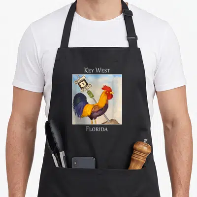 Mile rooster apron - Watercolor rooster beside Key West Begin North 1 Mile 0 Florida sign — shown on a black cotton canvas apron against a white background