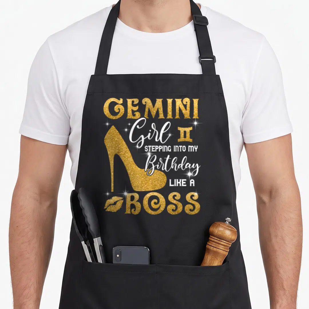 Gemini bbq boss apron - Gold glitter high heel with GEMINI Girl Stepping Into My Birthday Like A Boss text — shown on a black cotton canvas apron against a white background