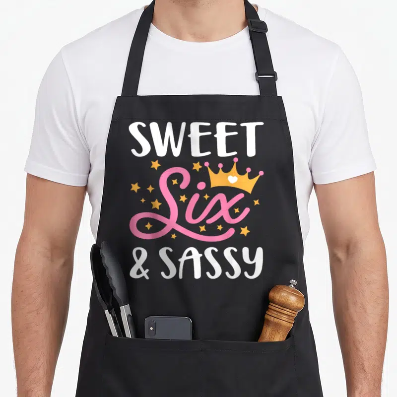 Six birthday sassy apron - Sweet Six Sassy text with orange crown and pink stars — shown on a black cotton canvas apron against a white background
