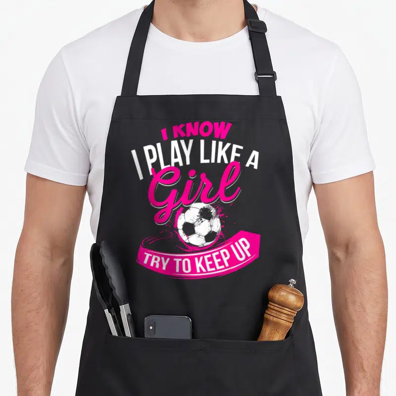 Funny play soccer apron - I Know I Play Like A Girl text with soccer ball and pink banner — shown on a black cotton canvas apron against a white background