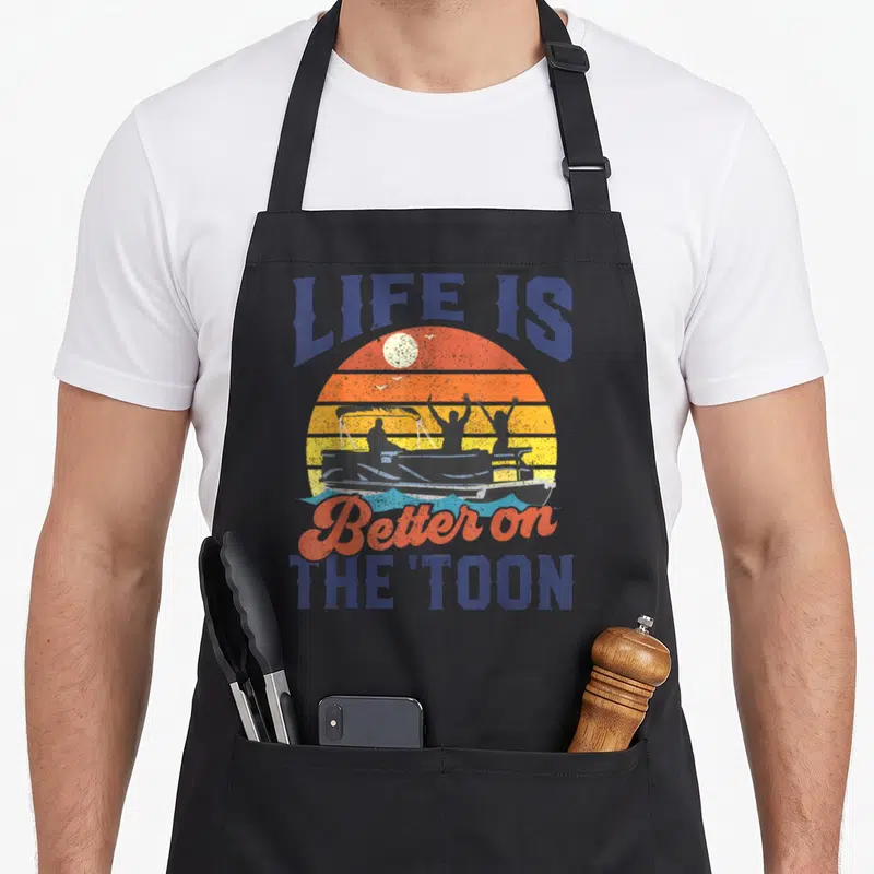 Better pontoon apron - Retro sunset graphic with pontoon boat silhouette and Life Is Better On The Toon text — shown on a black cotton canvas apron against a white background