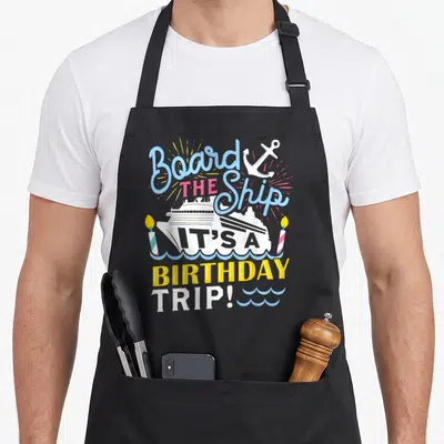 Cruise birthday apron - Typography design with ship, anchor, two candles, and waves in blue pink yellow white — shown on a black cotton canvas apron against a white background