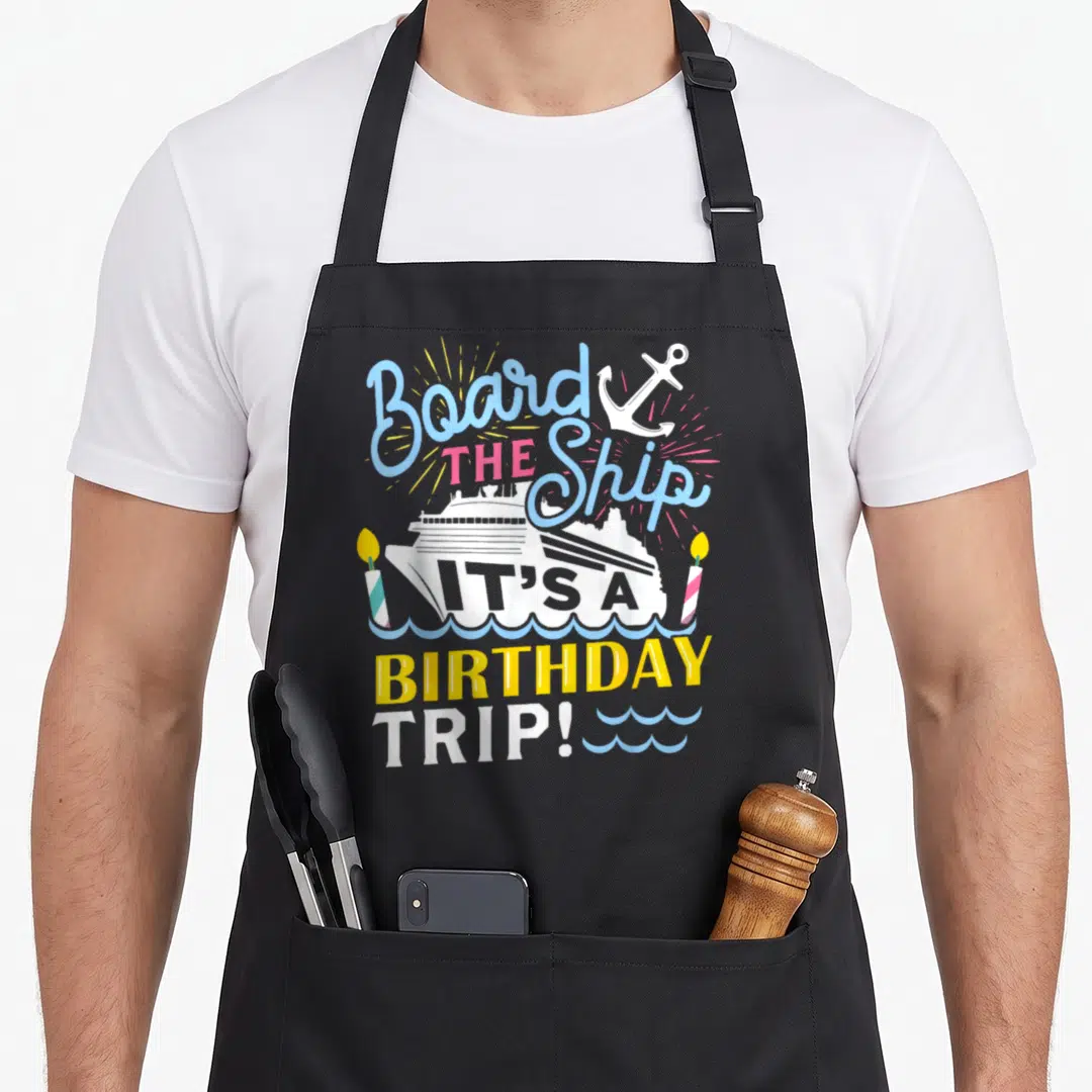 Cruise birthday apron - Typography design with ship, anchor, two candles, and waves in blue pink yellow white — shown on a black cotton canvas apron against a white background