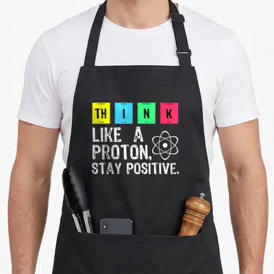 Proton stay science apron - Four colored element tiles spelling THINK above white atom symbol and Stay Positive slogan — shown on a black cotton canvas apron against a white background