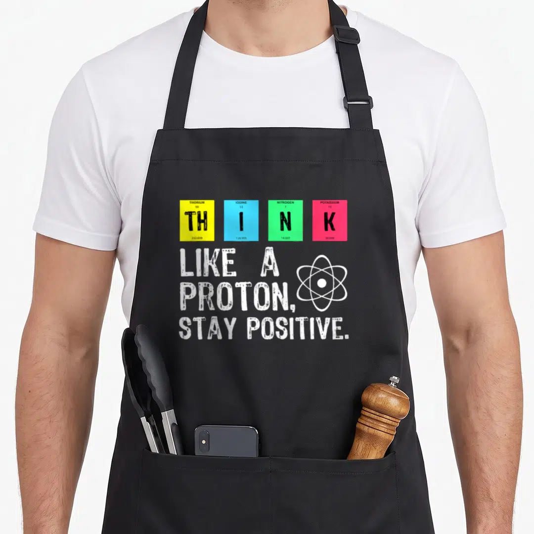 Proton stay science apron - Four colored element tiles spelling THINK above white atom symbol and Stay Positive slogan — shown on a black cotton canvas apron against a white background