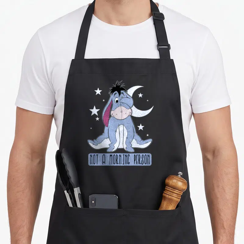 Funny morning eeyore apron - Eeyore sitting under moon and stars with NOT A MORNING PERSON text in blue and purple — shown on a black cotton canvas apron against a white background