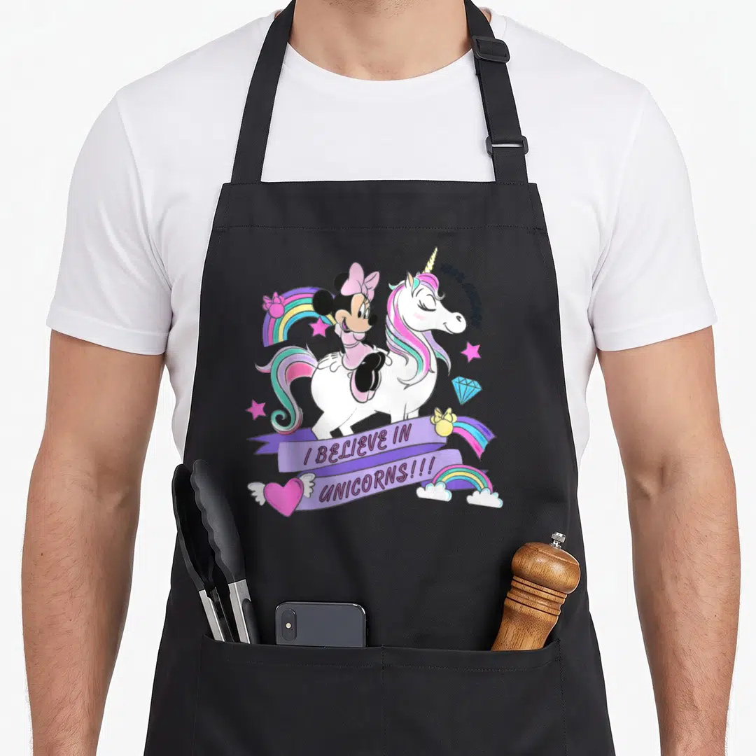 Cute unicorn apron - Minnie Mouse riding a white unicorn with pink mane and I believe in unicorns ribbon text — shown on a black cotton canvas apron against a white background
