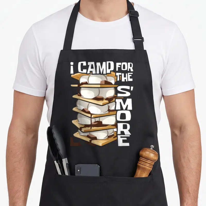 Smore campfire camping apron - Cartoon stack of five s'mores with white text reading i CAMP FOR THE S'MORE vertically — shown on a black cotton canvas apron against a white background