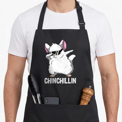 Dabbing pet apron - Cartoon chinchilla with sunglasses and pink ears dabbing above white text CHINCHILLIN — shown on a black cotton canvas apron against a white background