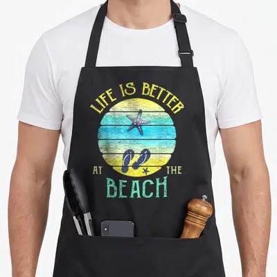Better beach life apron - Life is Better at the Beach text over striped sunset with starfish and flip flops — shown on a black cotton canvas apron against a white background